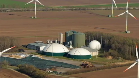 Biogazownia z białymi i szarymi zbiornikami oraz silosami między turbinami wiatrowymi w Land Wursten, widziana z samolotu.