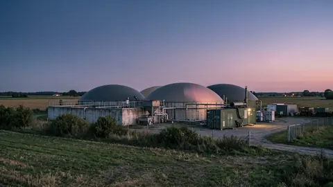 Biogazownia po zachodzie słońca: kopuły w ostatnim różowym świetle, ciepłe lampy przy kontenerze CHP i spokojne pole na pierwszym planie.