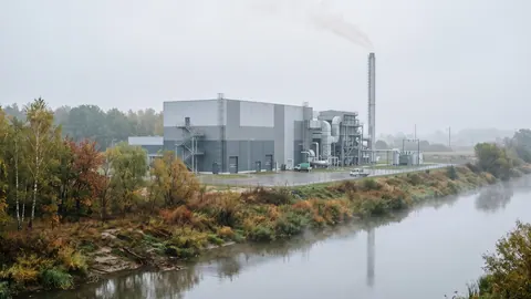 Nowoczesna instalacja termicznego przekształcania odpadów z wysokim kominem i halą kotłów nad rzeką, w mglisty jesienny poranek.