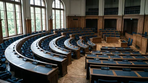 Pusta sala obrad plenarnych z półkolistymi rzędami drewnianych pulpitów i mikrofonów, w ciepłym świetle wpadającym przez wysokie okna.