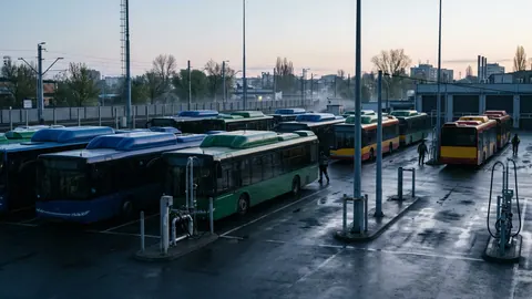Zajezdnia autobusowa o świcie: autobusy miejskie z zabudową butli CNG na dachach przy stanowiskach tankowania na mokrym placu.
