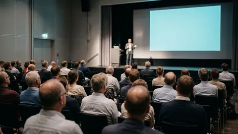 Uczestnicy konferencji słuchają prelegenta na scenie z pustym ekranem, widok zza publiczności w miękkim świetle.