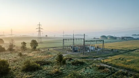 Linie wysokiego napięcia i stacja elektroenergetyczna o wschodzie słońca, w oddali biogazownia we mgle.