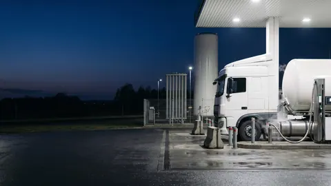 Nocna stacja bioLNG: ciągnik z kriogeniczną cysterną LNG przy dystrybutorze pod zadaszeniem, pionowy zbiornik LNG i odbicia świateł na mokrym asfalcie.