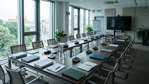 Pusta, nowoczesna sala posiedzeń rządowych z długim stołem, teczkami i raportami oraz widokiem na miasto przez duże okna.
