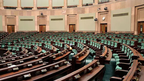 Pusta sala plenarna Sejmu RP w Warszawie: półkoliste rzędy drewnianych ław poselskich i zielonych foteli.