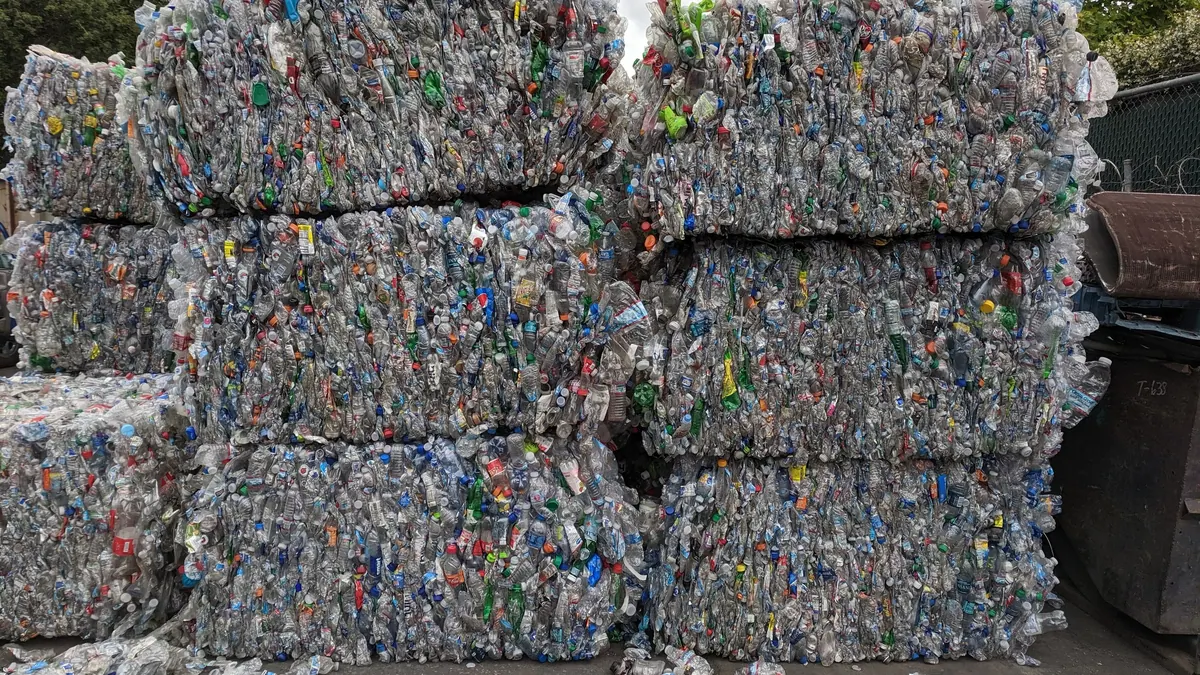 Sprasowane bele zgniecionych butelek PET ułożone w stosy na placu zakładu recyklingu, pod zachmurzonym niebem.