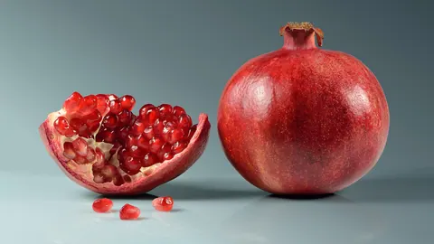 Cały owoc granatu i rozłamany owoc z odsłoniętymi, rubinowymi osnówkami nasion na szaroniebieskim tle.