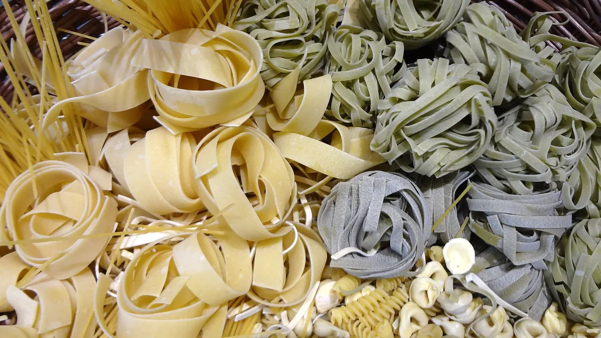 Różne rodzaje makaronu – świeże wstążki tagliatelle, makaron szpinakowy, spaghetti i muszelki – rozsypane obok siebie.