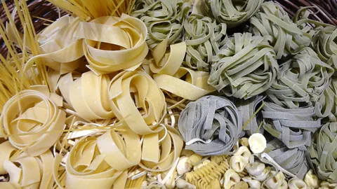 Różne rodzaje makaronu – świeże wstążki tagliatelle, makaron szpinakowy, spaghetti i muszelki – rozsypane obok siebie.