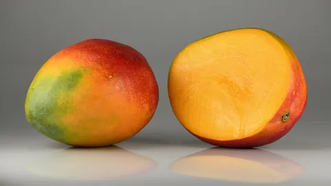 Dojrzałe mango z czerwono-zieloną skórką w całości oraz przekrojone na pół z widocznym pomarańczowym miąższem.